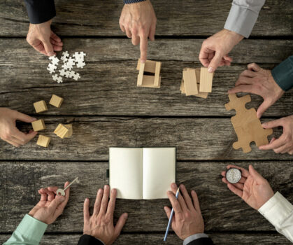 Eight businessmen planning a strategy in business advancement each holding  different but equally important metaphorical element - compass,  puzzle pieces, pegs, cubes, key and one making notes.